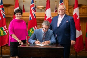 MPP Kevin Holland signing the oath of office in Queen's Park, with Premier Doug Ford and Secretary of the Cabinet Michelle E. DiEmanuele at his side. The image captures a crucial step in his political career, underscoring the solemnity and responsibility of his new role.