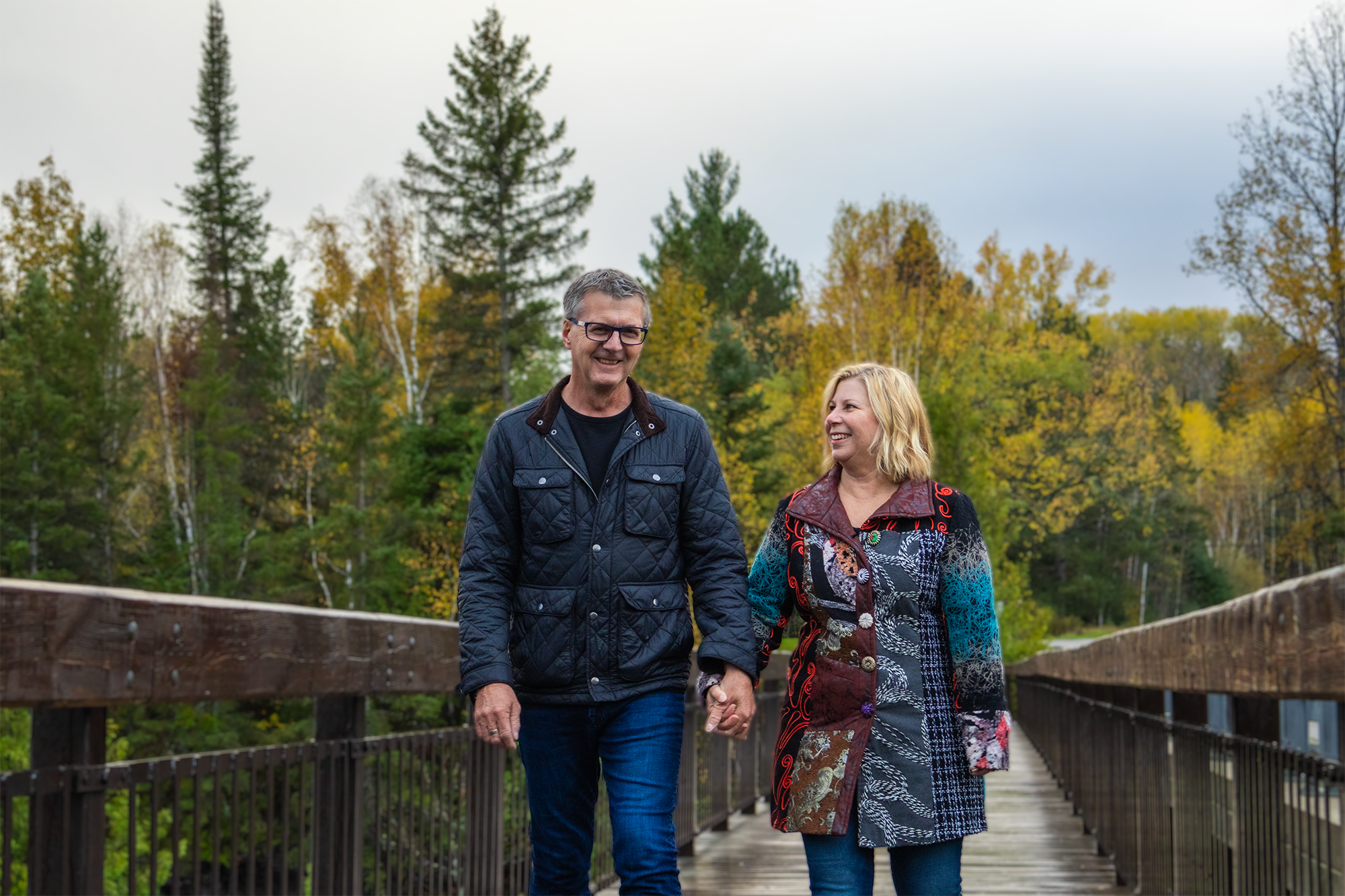 MPP Kevin Holland and his wife Lori walking on a bridge, with Lori looking up at Kevin, who is looking forward. The background is adorned with trees showcasing autumn colors, adding a serene and picturesque quality to the scene.