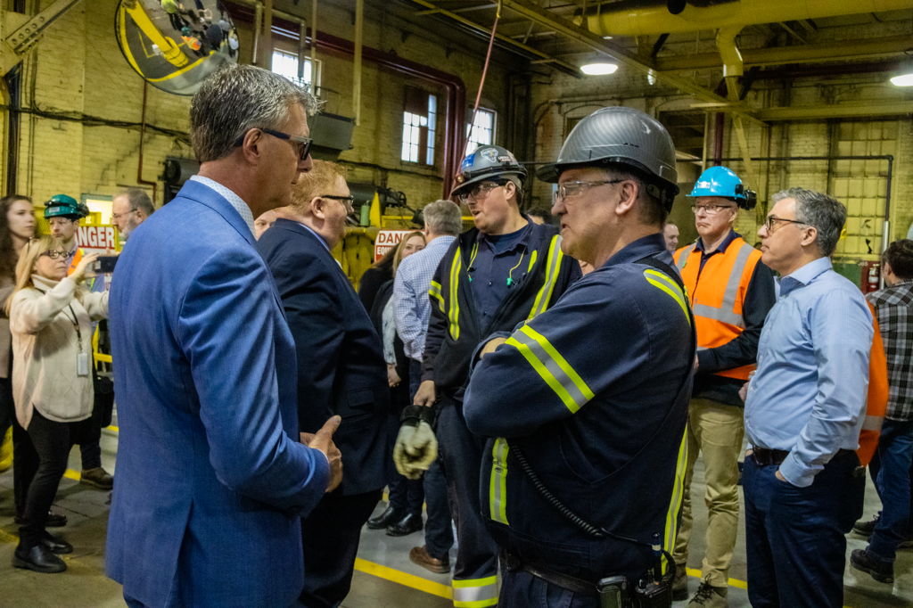 MPP Kevin Holland engaged in conversation with mill workers after an MNRF announcement. The image shows MPP Holland dressed in business attire, speaking with a group of workers who appear attentive and engaged.